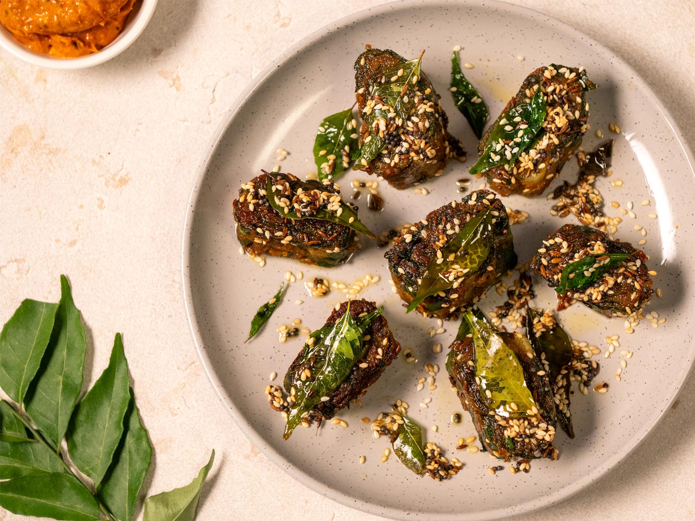 A white plate containing 7 oval-shaped pieces of patra -- crispy fried segments of leaves layered with spiced chickpea flour -- sprinkled with seeds and herbs.