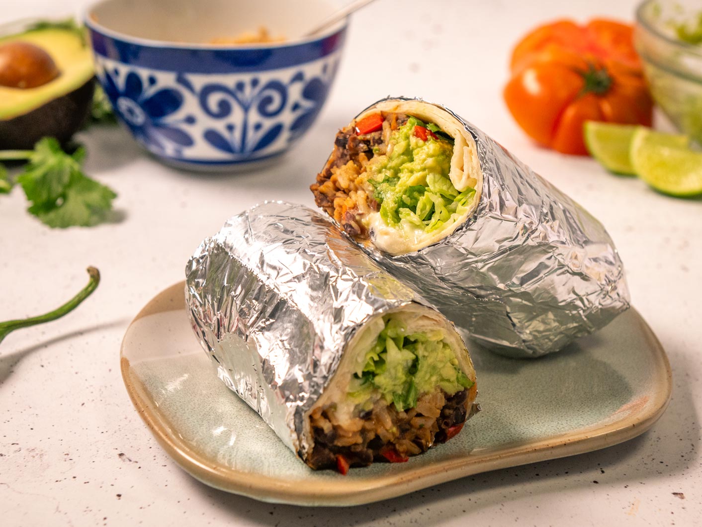 A hearty looking burrito wrapped in foil and sliced in half, revealing the refried bean mixture, avocado, lettuce, and various other fillings inside.