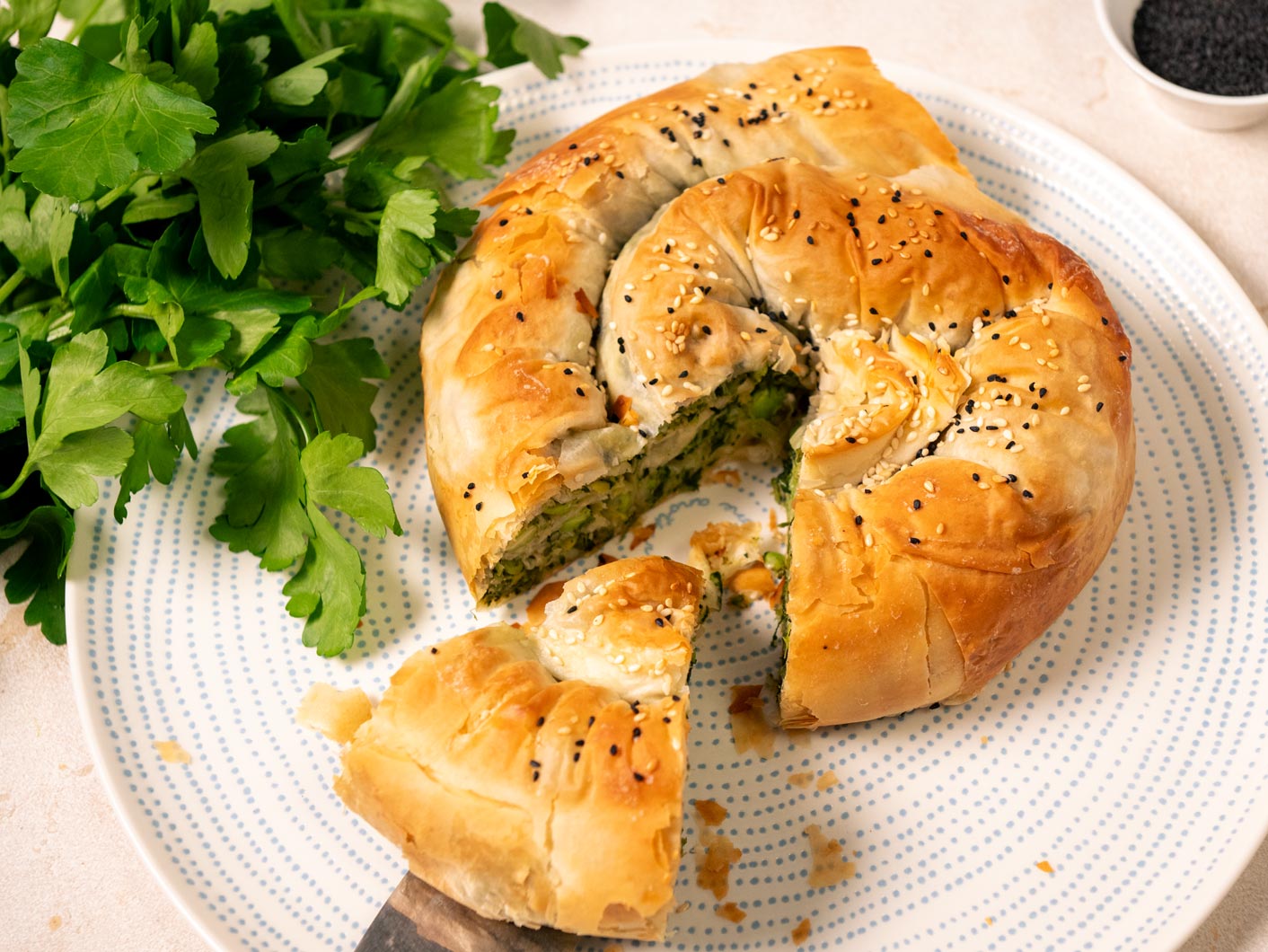 A spanakopita sits on a light coloured plate while a serving utensil pulls out a slice, revealing the inside.