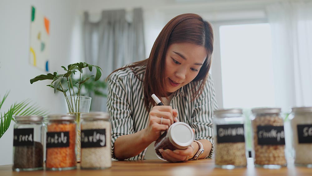 A woman writes a label on a label on a food storage jar, while other labelled jars sit blurry in the foreground. 