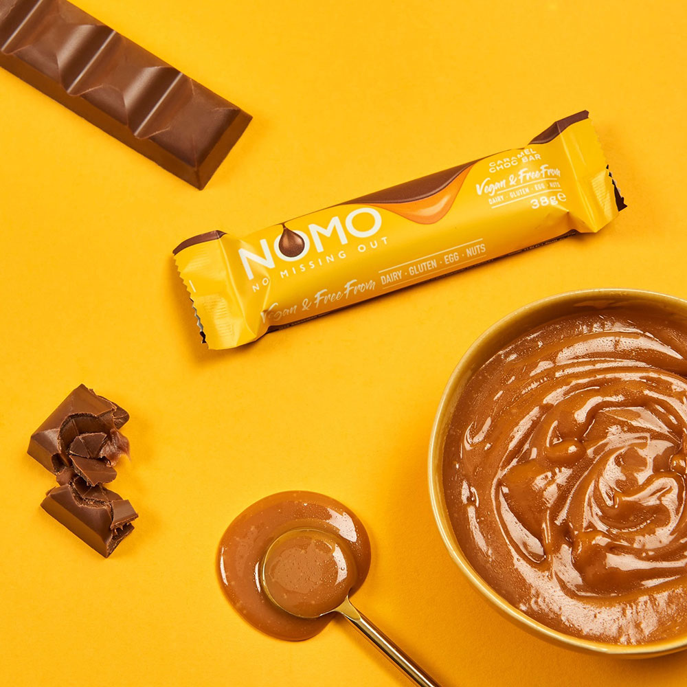 A bar of NOMO's caramel sea salt is on a bright yellow background. A bowl of creamy caramel sits off to the side.