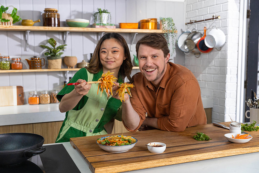 Simon Toohey and Liz Miu behind the bench of the Freshly Picked kitchen., holding up a delicious bowl of food.