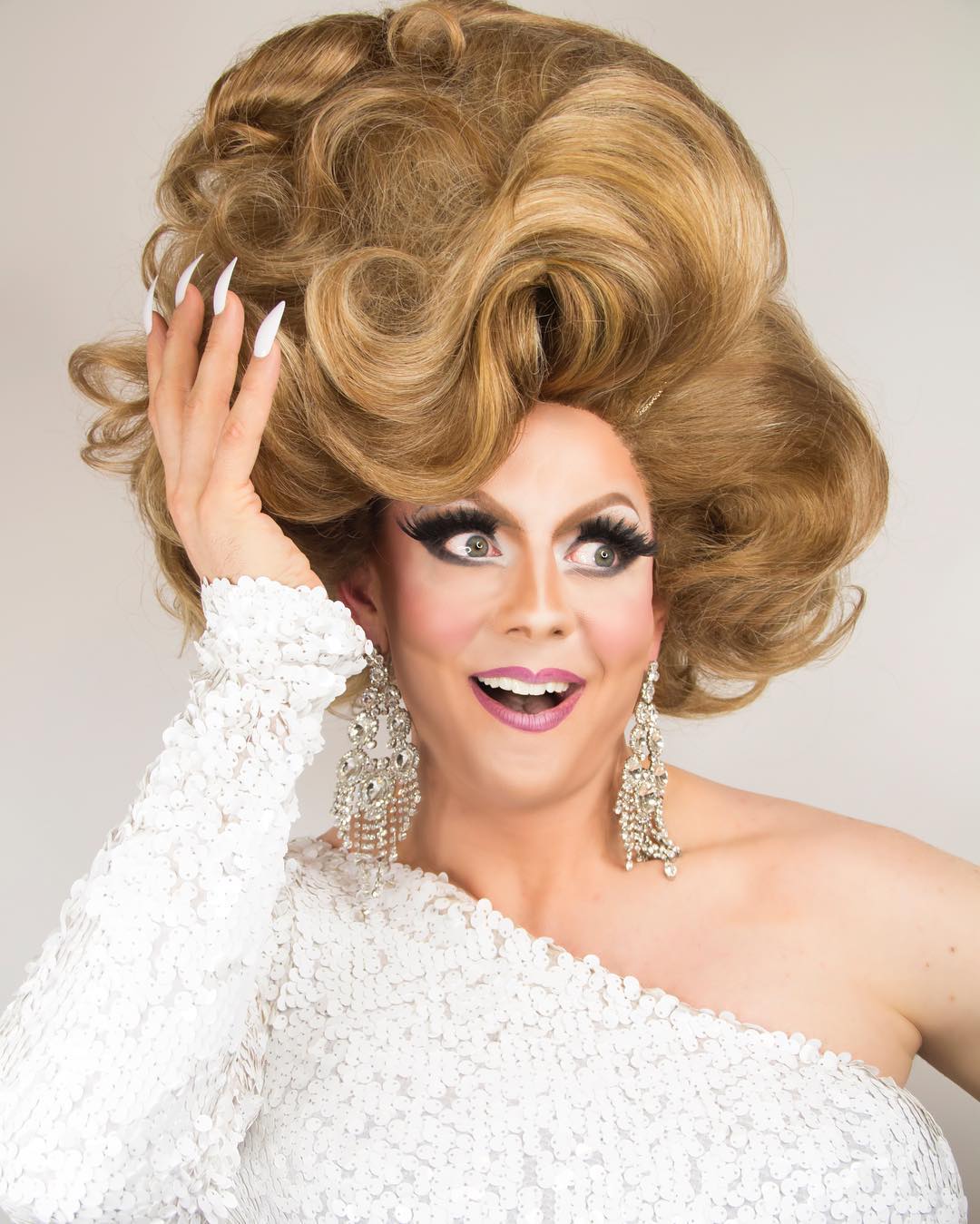 A close-up portrait of Honey La Bronx wearing a high brunette wig and gown reminiscent of 1920s Hollywood. She is smiling and holding her hair.