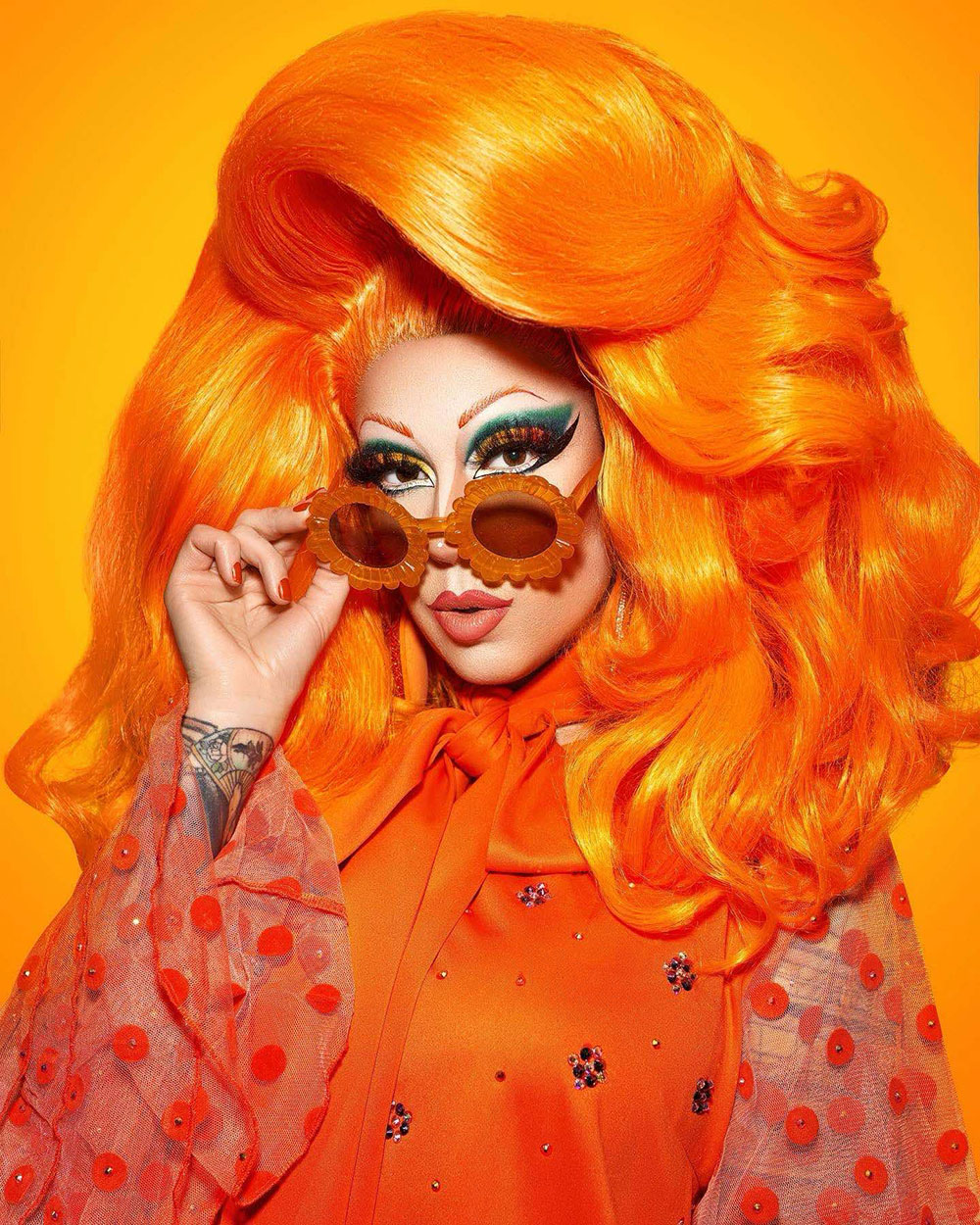 Mardi Gras 2025 - Fay stands in front of an orange background wearing a tall orange wig with her hair down, a vintage orange dress with lace sleeves and a pussy bow, and holds her orange 'flower power' sunglasses just off her nose, revealing exquisitely painted eye makeup.