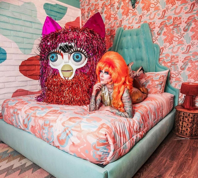 Fay Ludes laying on a bed on her stomach, facing the camera. She is wearing her signature orange with vintage styling, while an oversized furby toy sits next to her.