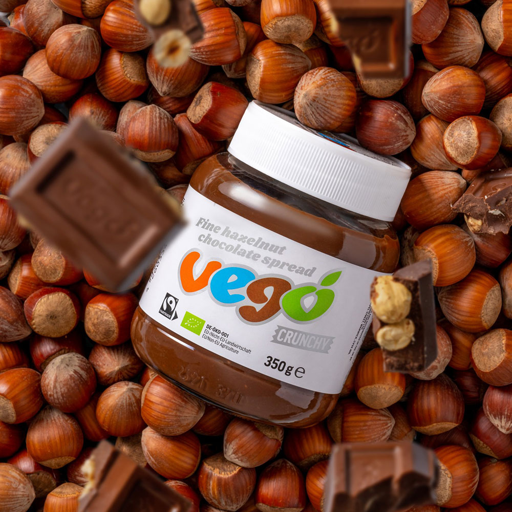 A jar of Vego choc hazelnut spread sits among pieces of chocolate and whole hazelnuts.