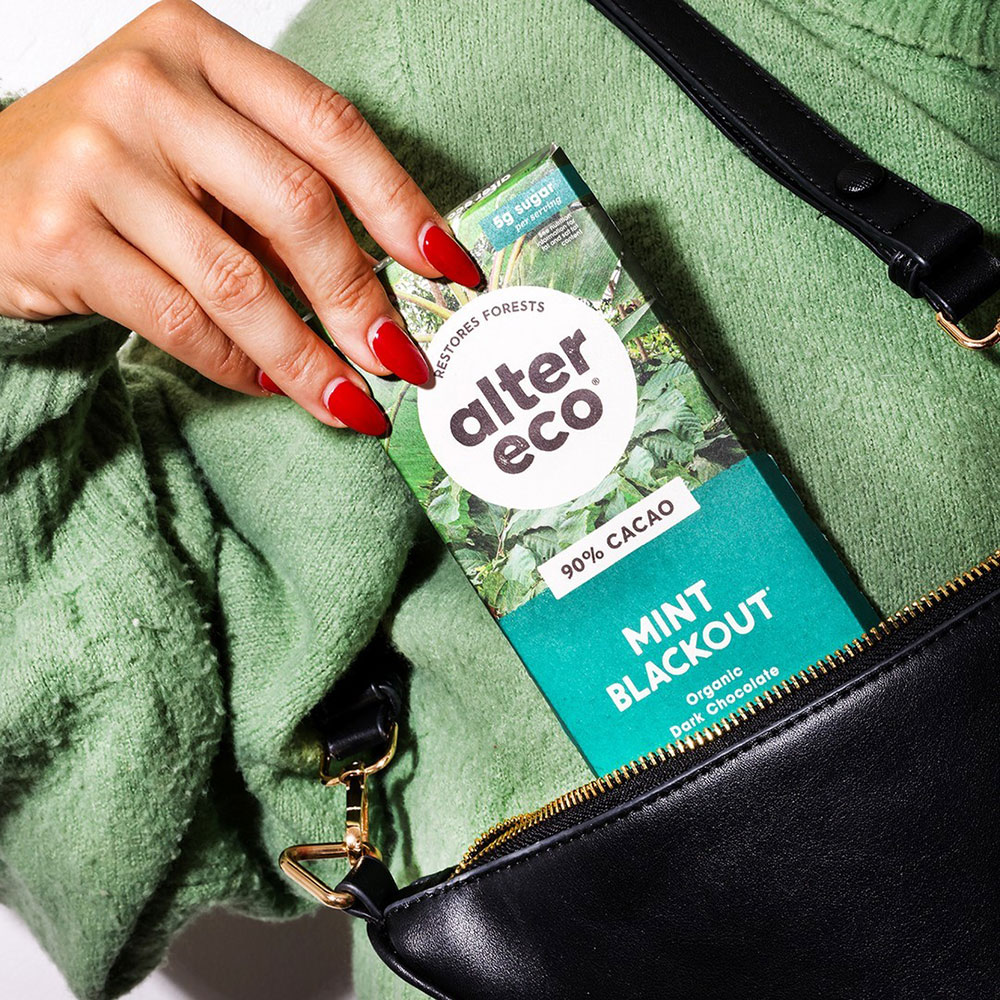 A hand is lowering a block of Alter Eco Mint Blackout chocolate into a handbag.