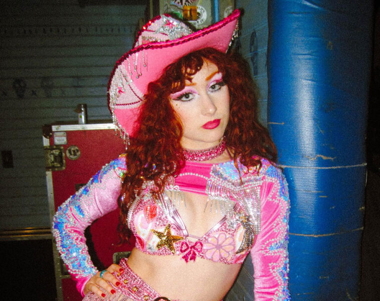 Chappell Roan faces the camera, hand on hip, wearing a pink cowboy hat and pink, purple and gold cropped top.