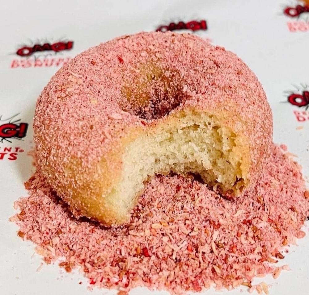 A close-up of a donut covered in pink sugar. It has a bite taken out and sits on a mountain of more pink sugar. 