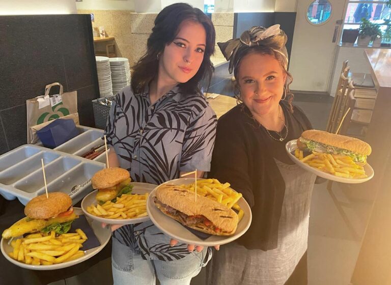 Two wait staff hold plates featurinmg a range of traditional pub fare, including burgers and fries.