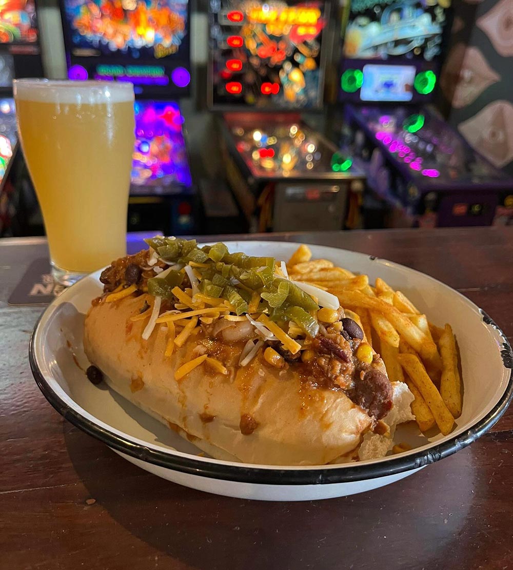 A vegan chilli cheese dog and a beer on a dining table, with pinball machines in the background.