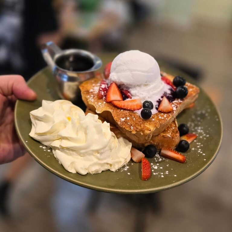 A hand holds a plate of vegan waffles with ice cream, berries, whipped cream and a side of maple syrup.