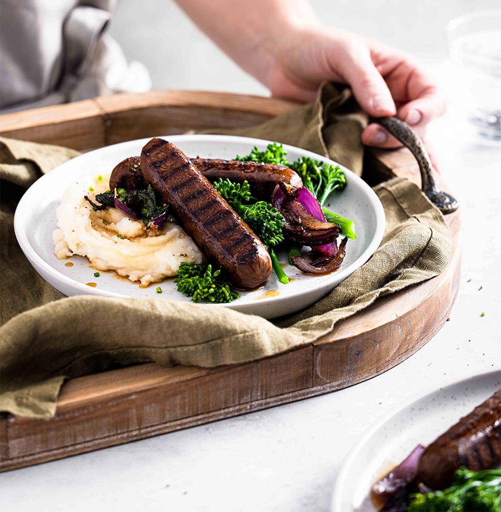 A person carries a tray holding a plate of Vegie Delights sausages served with mash and greens.