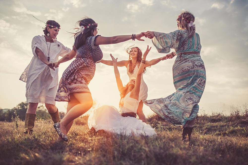 A group of women in flowy dresses dance around a seated woman with her arms in the air. They're in a meadow, and some of the women are wearing flowers in their hair. 