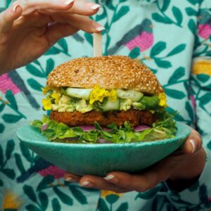 A person holds a green goddess burger from Yumi's on a turquoise plate. The person is wearing a patterned turquoise top with birds on it.