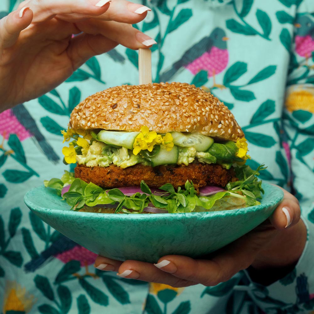 A person holds a green goddess burger from Yumi's on a turquoise plate. The person is wearing a patterned turquoise top with birds on it.