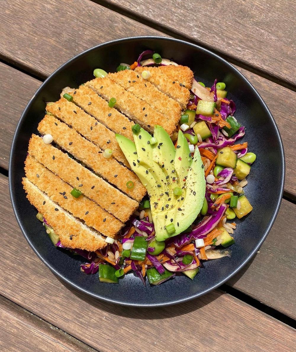 A vegan chicken katsu is presented in a black bowl on a wooden bench.