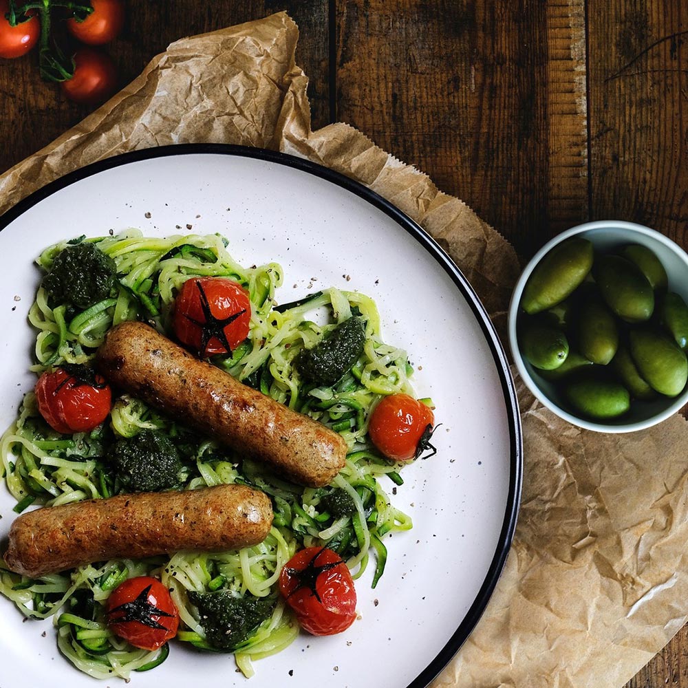 An overhead view of a plate of greens, tomatoes, and Unreal Co vegan sausages. A ramekin of cornichons sits to the side. 