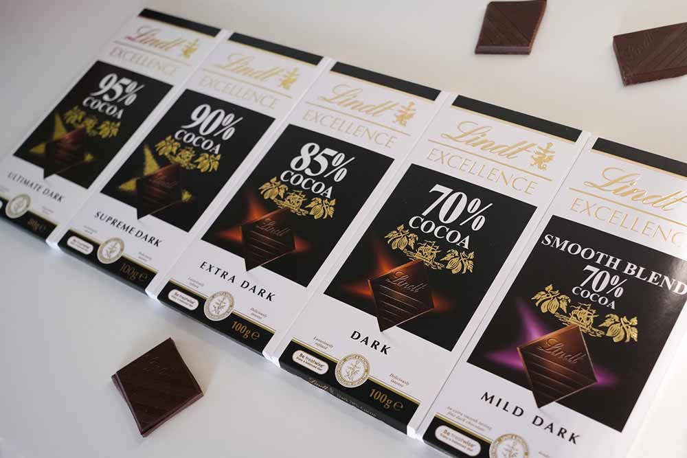 Six Lindt Excellence dark chocolate bars with different cocoa percentages, lined up next to each other on a white surface, surrounded by four pieces of plant-based chocolate.