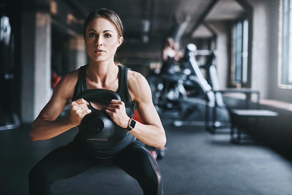 A woman in a gym is doing squats while holding a kettle bell. 