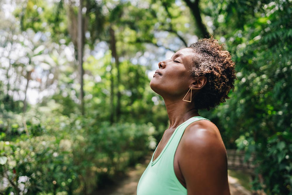 A woman out in nature looking calm with eyes closed, taking a deep breath.