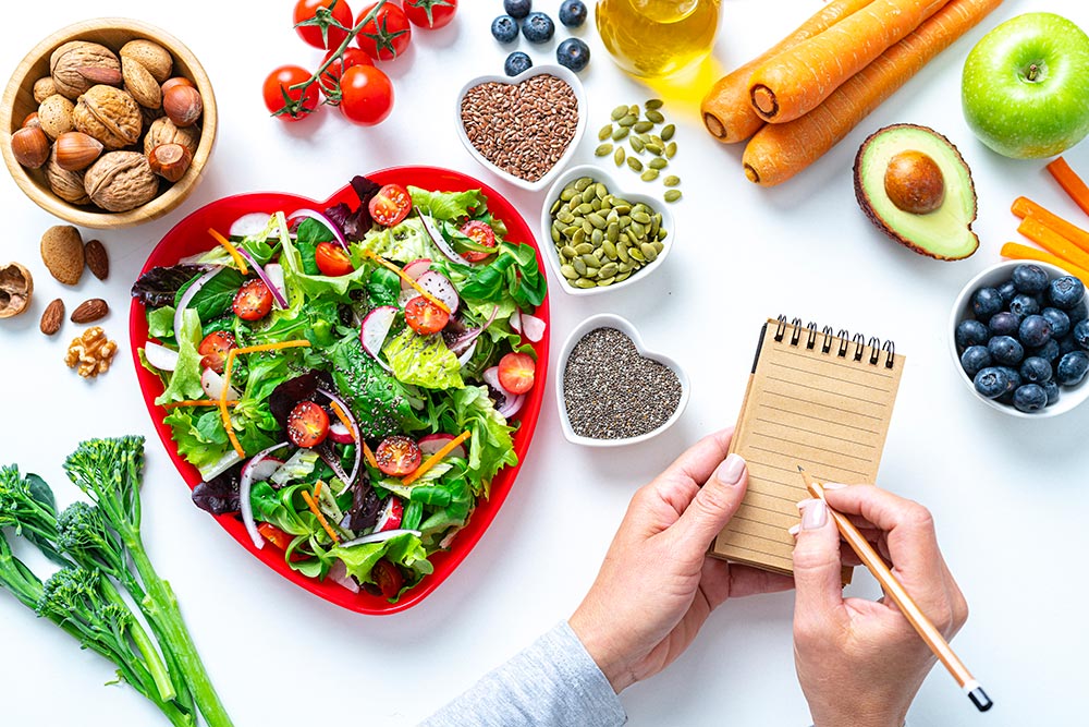 Overhead view of a red heart shape plate with healthy salad shot on white background. Female hands are writing with a pencil on a note pad. Multi colored fresh fruits,