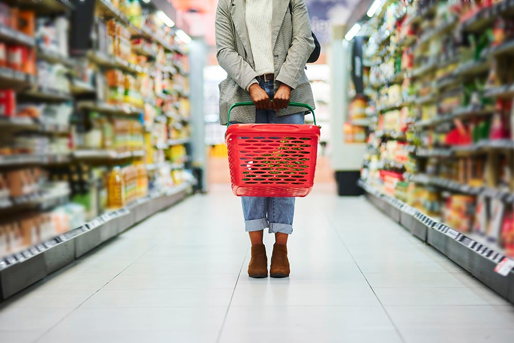 A shoulders-down shot of a person standing in the middle of a supermarket aisle holding a basket down at their knees. 