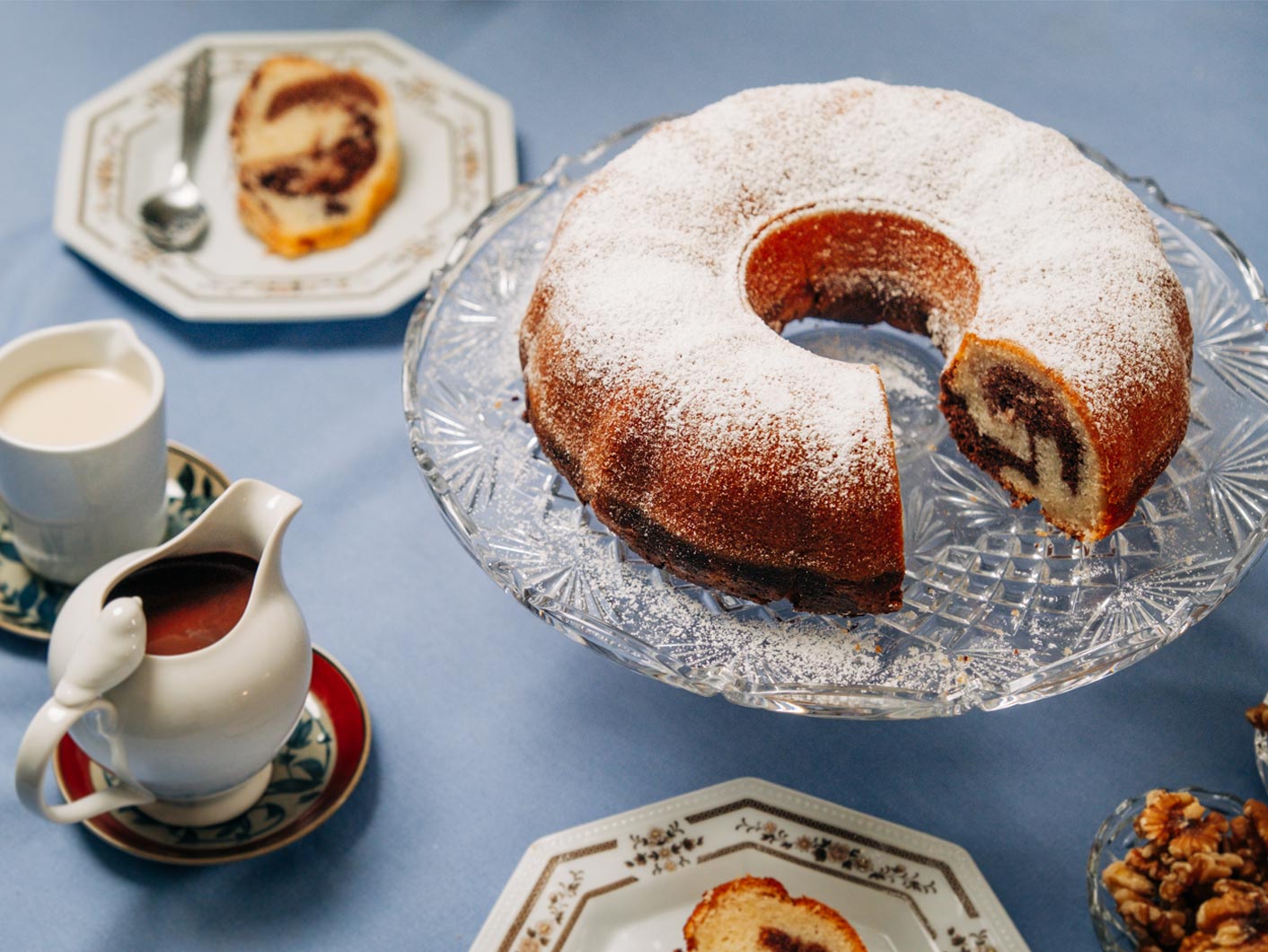 A marble bundt cake sits atop a glass cake stand, with a blue tablecloth underneath.