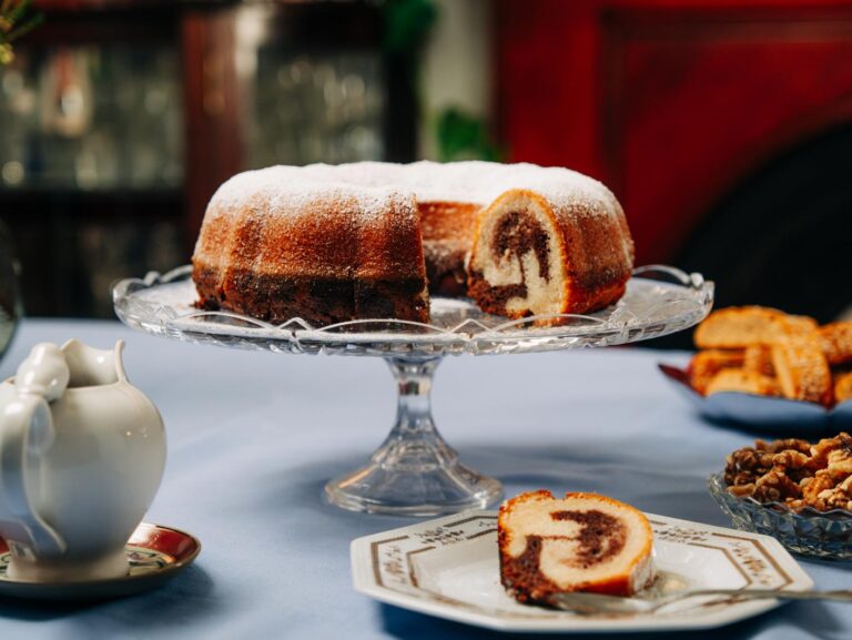 A marble bundt cake sits atop a cake stand, dusted with icing sugar. A slice has been cut and served on a small plate, revealing the marbled centre.