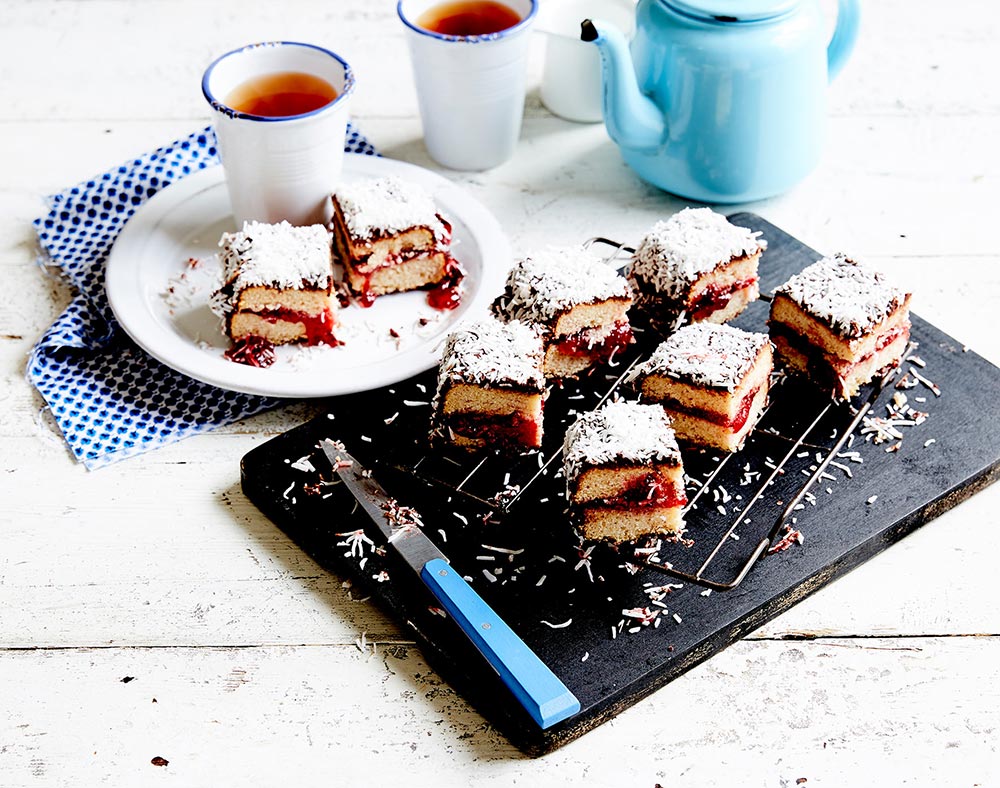 A tray of individually portioned lamington slice, filled with sweet jam. Cups of tea and a teapot sit in shot. 