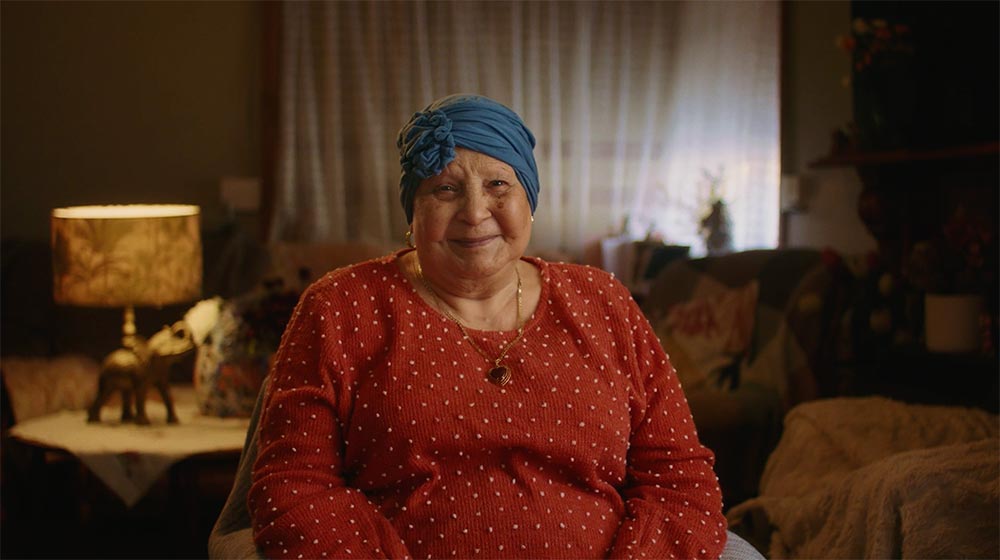 Nanna Mirella sits on the couch in her lounge rooms, smiling to camera. 