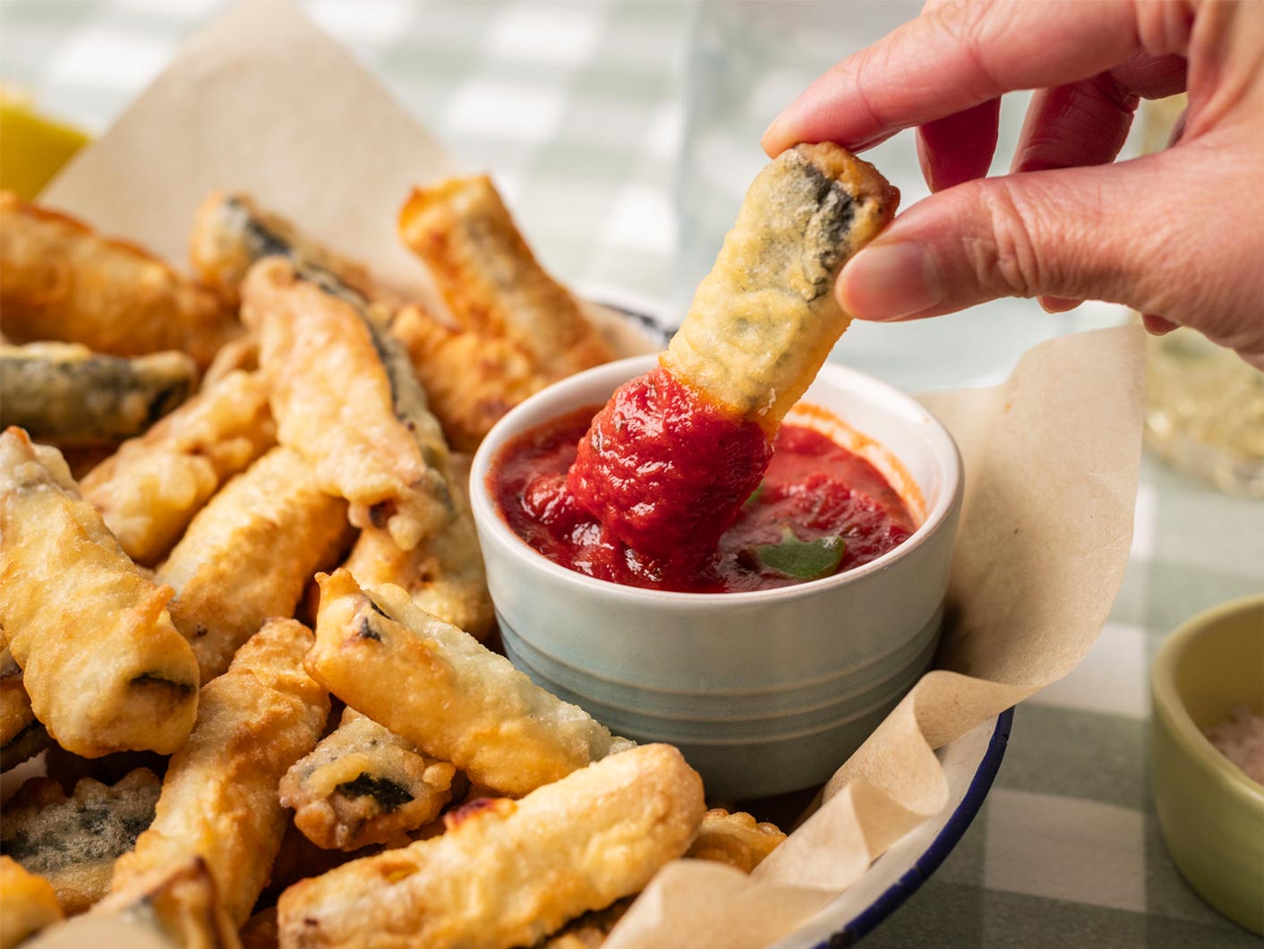 A hand holds a zucchini chip from the bowl and dips it into Napoli sauce