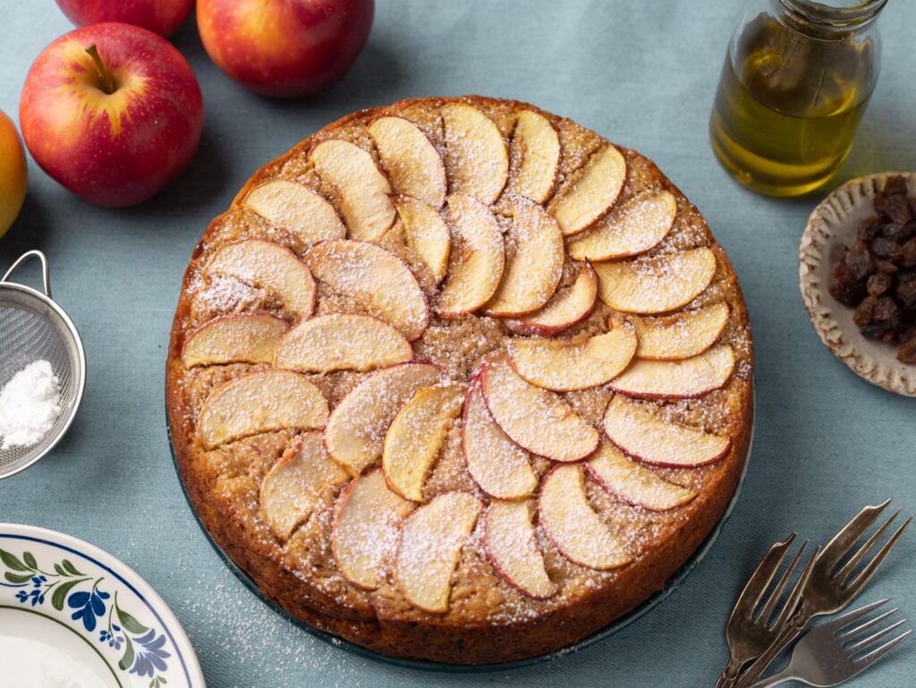 A beautifully decorated apple cake sits on a blue bench top. It is garnished with thin half-moon apple slices and dusted with icing sugar.