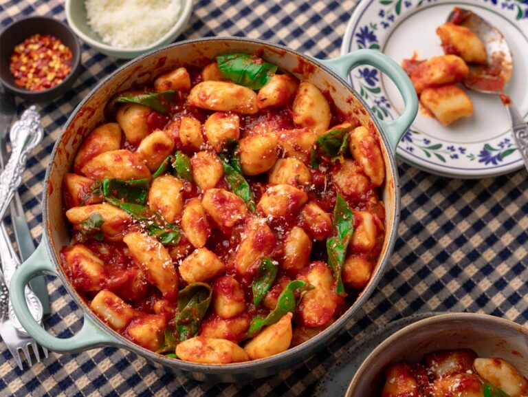 A serving bowl full of gnocchi in a Napoli sauce sits on a checked black and white table cloth.