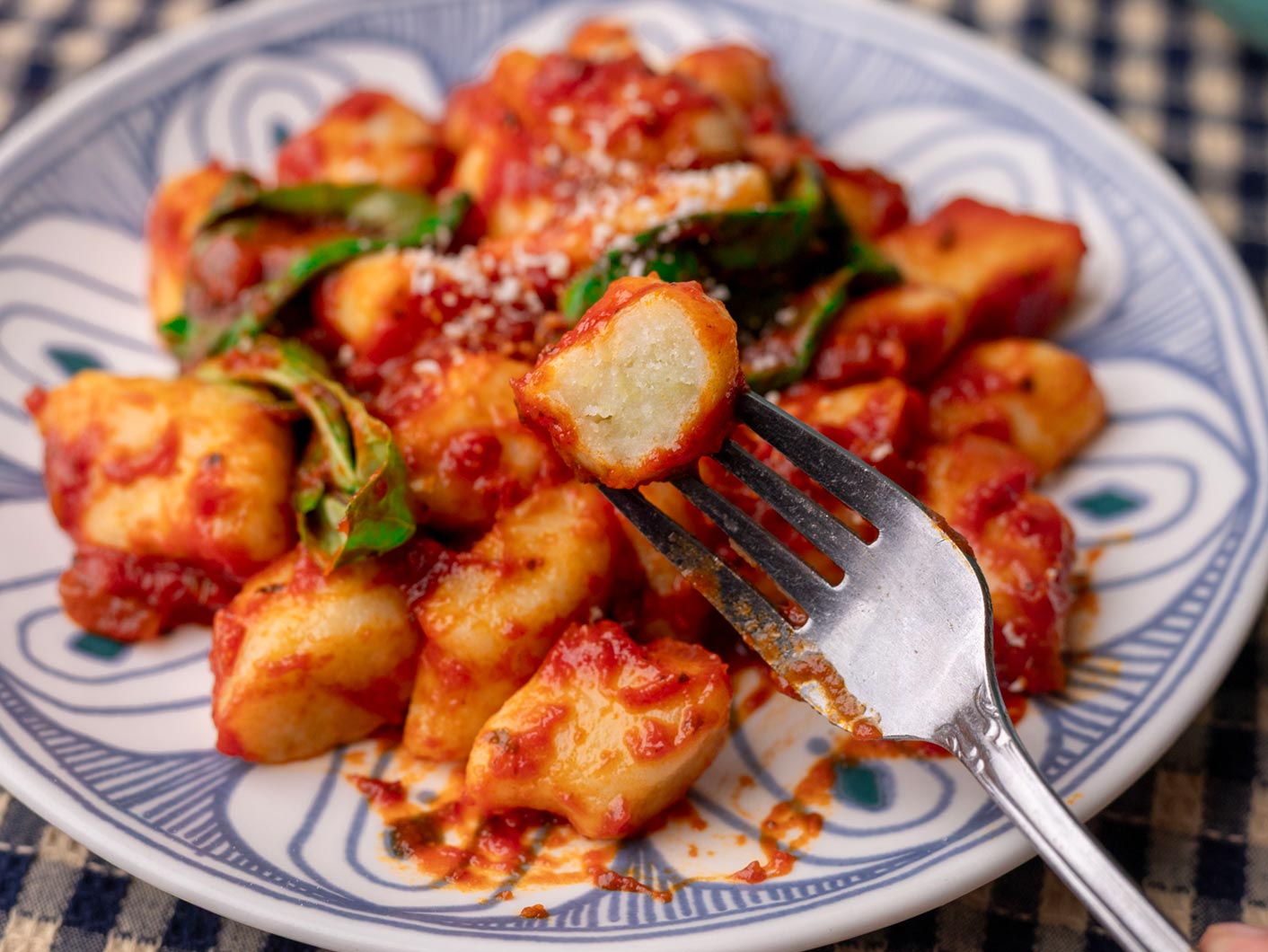 A plate of gnocchi with a fork holding a single potato dumpling to camera with a bite taken out, revealing it's pillowy centre.