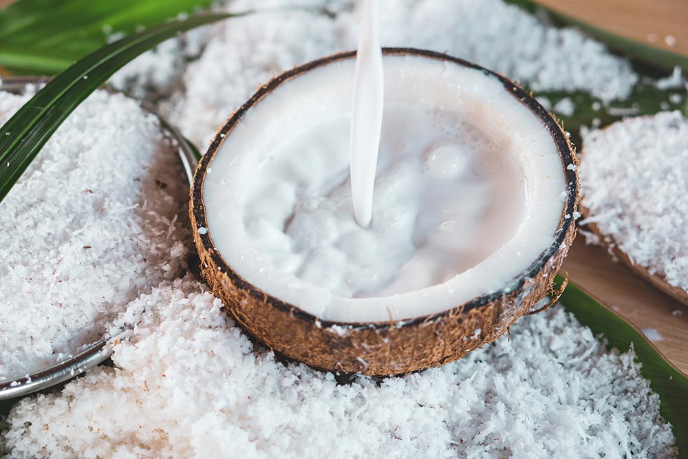 A halved coconut full of milk sits on a bench covered in desicated coconut. 