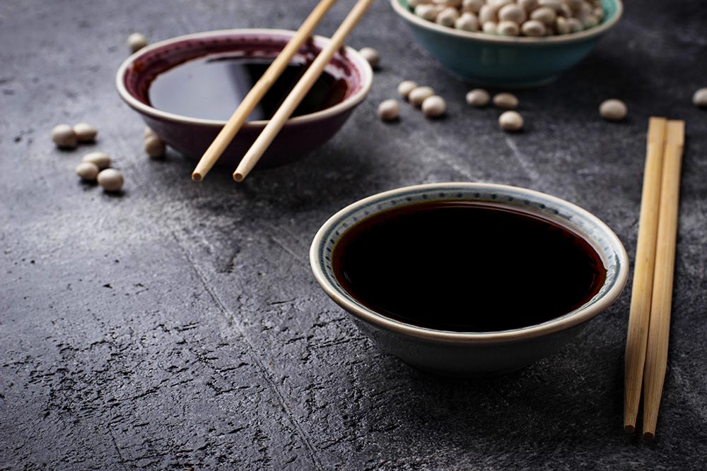 Two small ceramic condiment dishes of teriyaki sauce. A pair of chopsticks balance on one dish while another pair sit beside the other. 