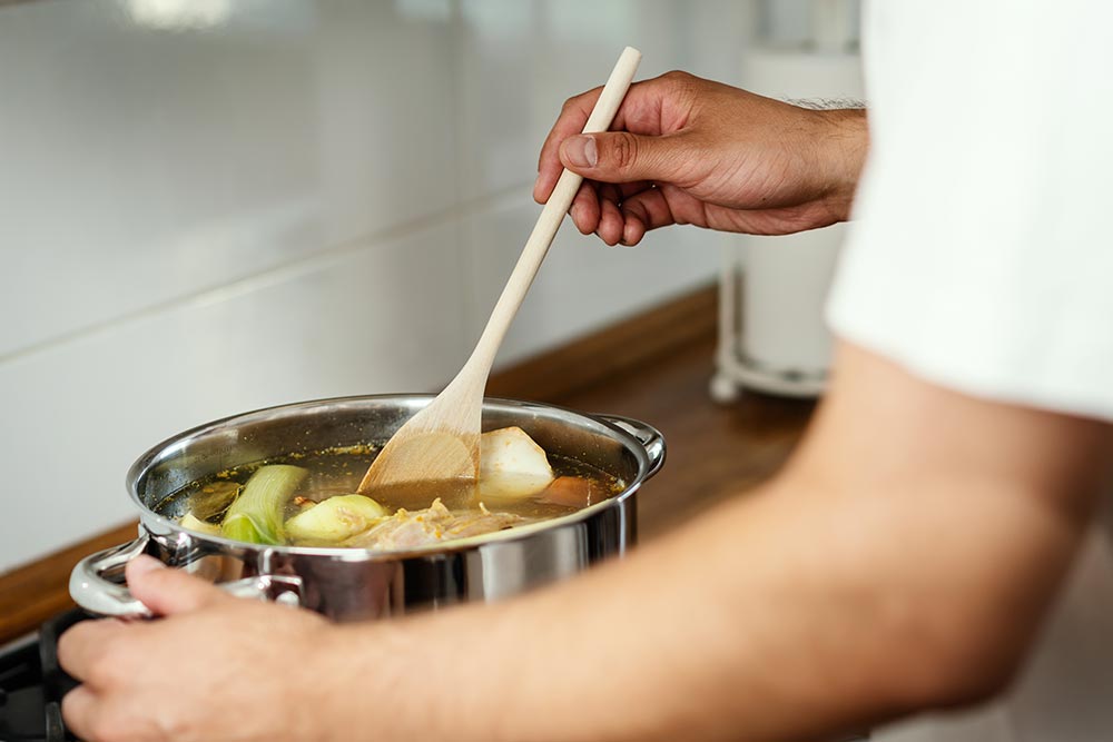 A person in a white t-shirt stirs a pot of vegetables on the heat, making a stock. 