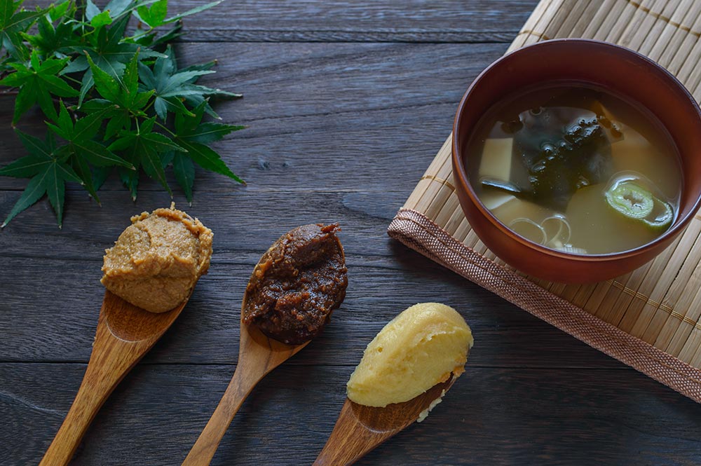 Three wooden spoons sit on a bench next to a small bowl of kombu. All three spoons have different kinds of miso paste on them - secret weapon’ vegan seasonings