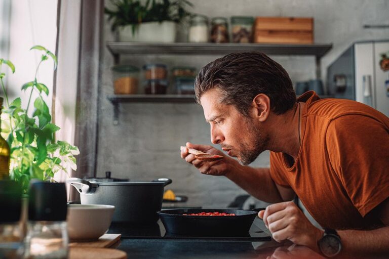 A person in an orange top leans over a pan on the stove and tastes a little of what they're cooking.