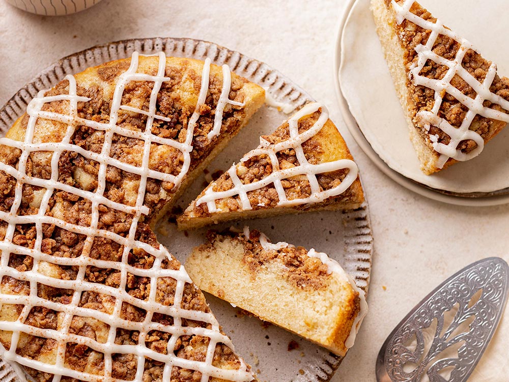 A cinnamon crumble tea cake sits on a plate with some slices cut out and in view. There is a lattice of white icing on top. 