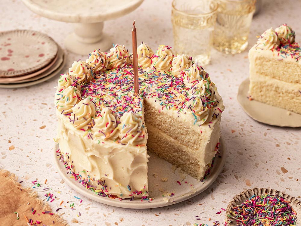 A beautifully decorated two-tiered vanilla birthday cake, with a buttercream petal design on the outside, and loaded with colourful sprinkles on top. A single candle is placed in the middle. 