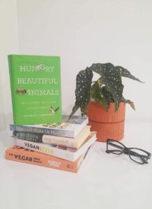A stack of books sit on a table next to a plant and some reading glasses.