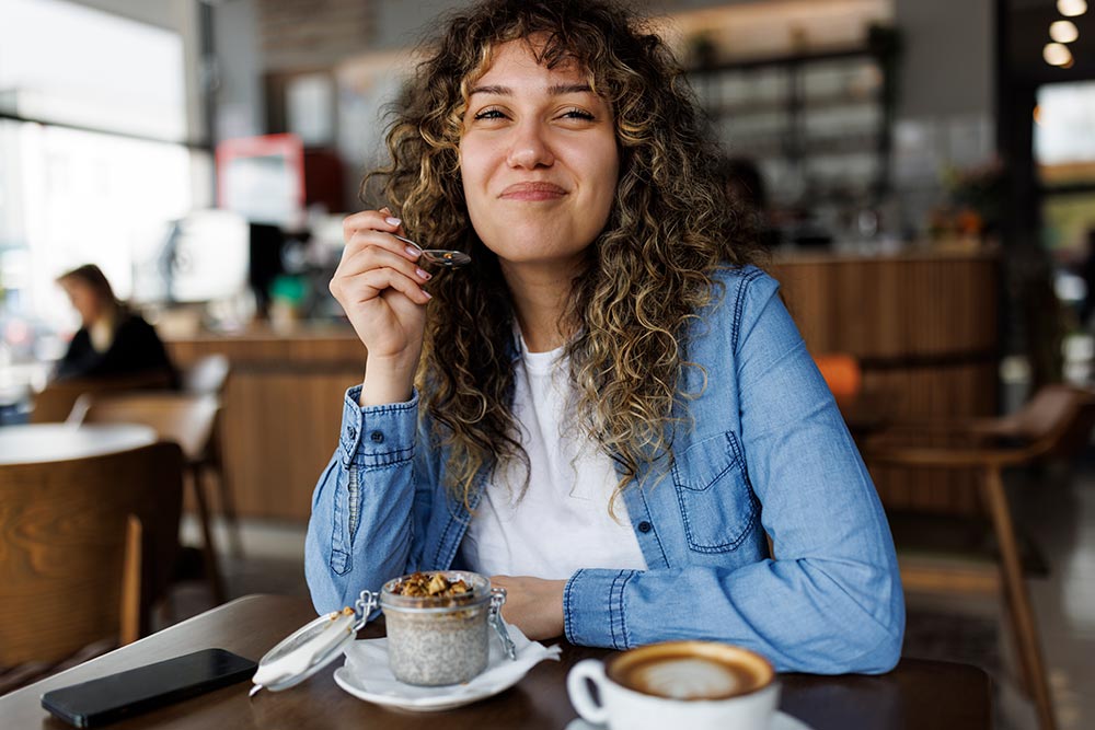 A woman sits in a cafe smiling while enjoying a coffee and chia pudding.