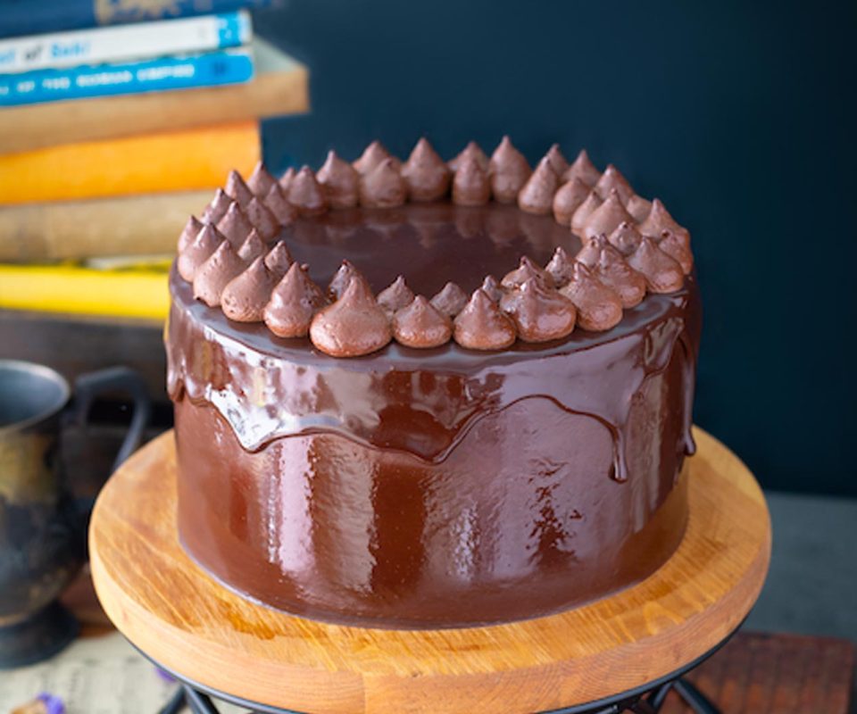 A round plant-based chocolate cake with glossy vegan ganache and piped dairy-free chocolate icing sits on a wooden cake stand.