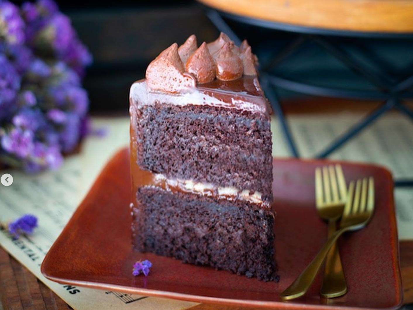 A slice of layered plant-based chocolate cake with vegan chocolate icing sits on a brown plate next to two gold forks, with purple flowers and a sheet of music in the background.