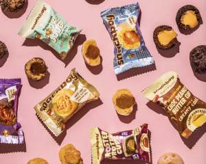 Assorted Snackboy packaged treats and opened mini desserts, featuring plant-based options, are arranged on a pink background, displaying a variety of flavours and fillings.
