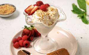 Two scoops of dairy-free ice cream topped with nuts, strawberries, and raspberries in a glass dish on a plate with bikkies.