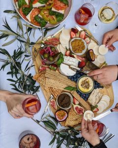 A plant-based cheese platter with biscuits, fruit, olives, and spreads, surrounded by hands holding glasses.