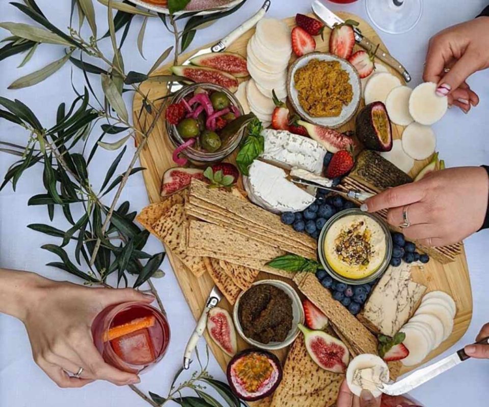 A plant-based cheese platter with biscuits, fruit, olives, and spreads, surrounded by hands holding glasses.